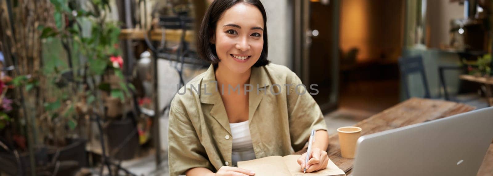 Portrait of young asian woman working on laptop, making notes, writing down while attending online lesson, work meeting by Benzoix