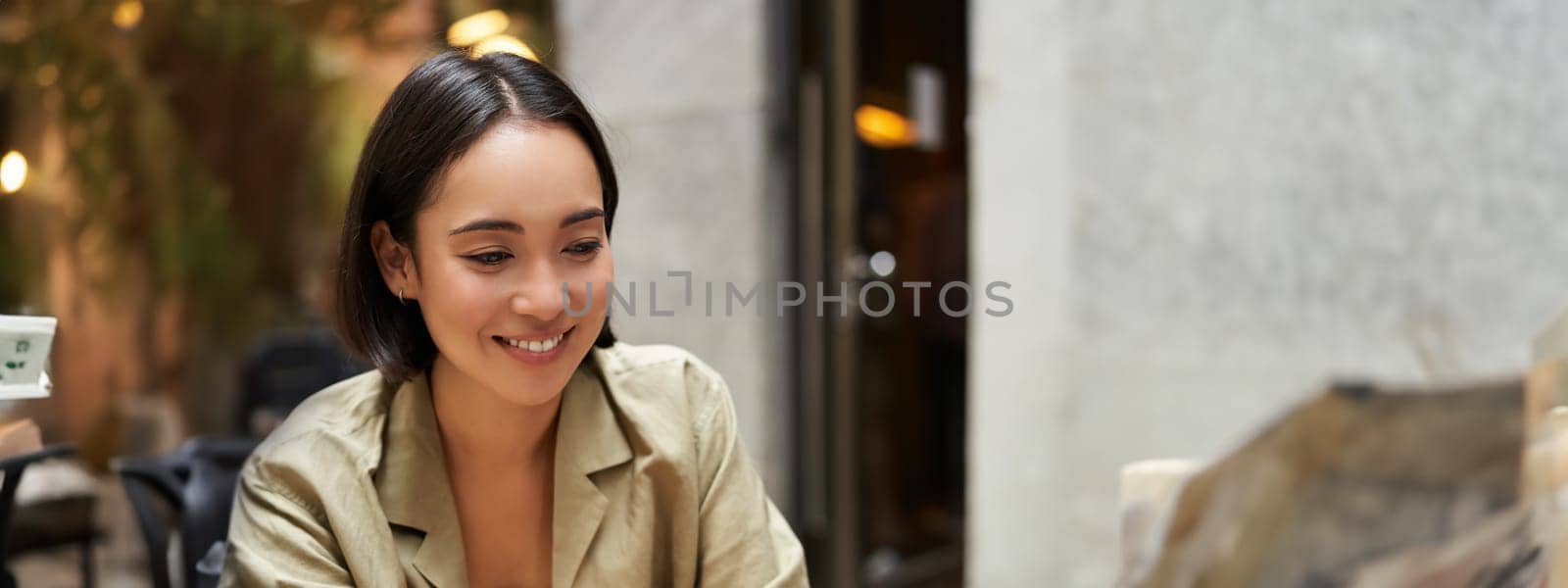 Vertical shot of young asian woman working on remote from outdoor cafe, sitting with laptop and smiling, studying.