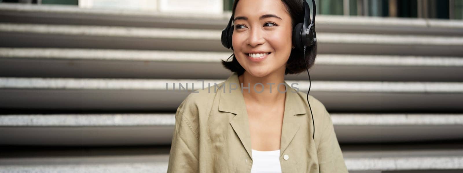 Portrait of beautiful girl in earphones, listening music, smiling and walking in city centre by Benzoix