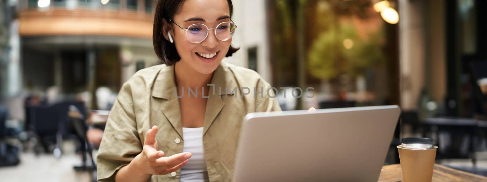 Online meeting. Young urban girl, asian woman talking with laptop, video chat, gesturing, sitting in an outdoor cafe and working remotely.