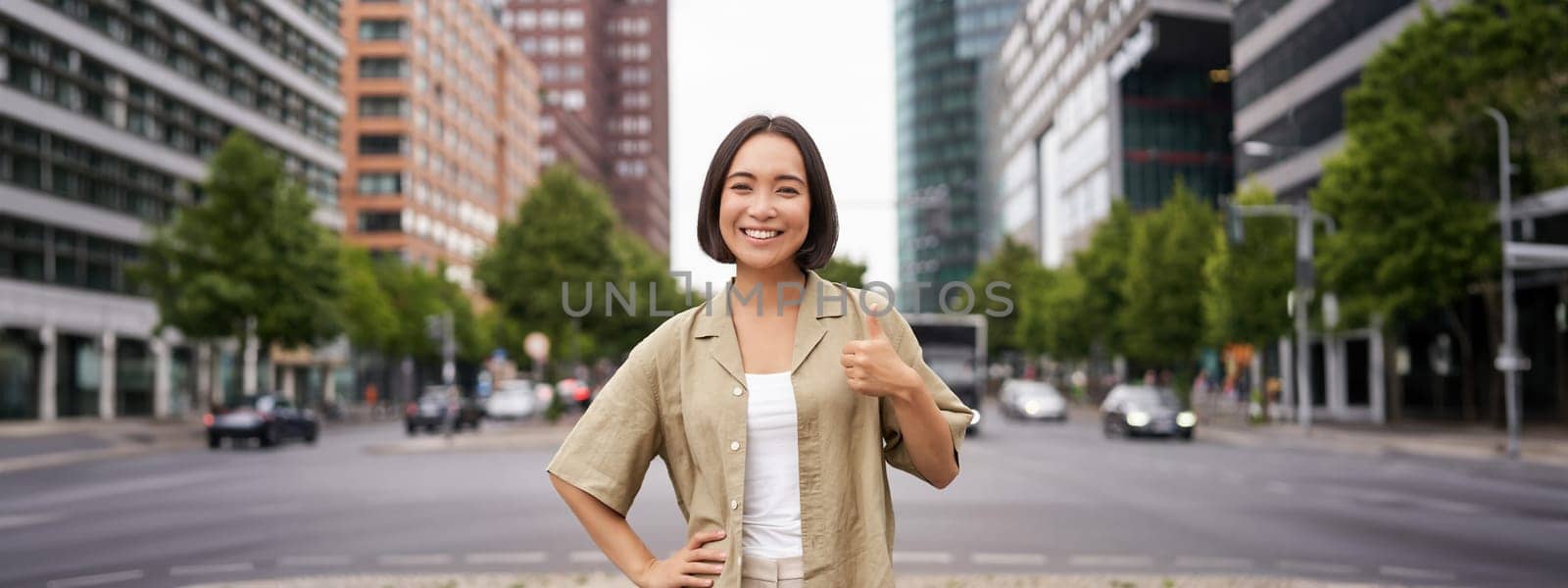 Enthusiastic city girl, shows thumbs up in approval, looking upbeat, say yes, approves and agrees, stands on street.