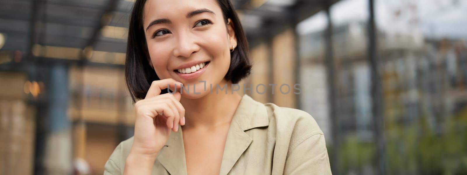 Close up portrait of asian girl with happy face, smiling and laughing, touching her skin, standing on street on sunny day.