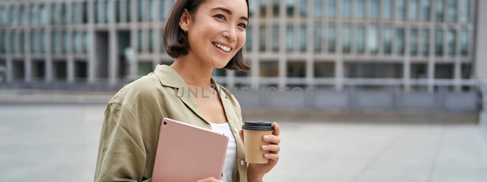 Portrait of happy asian girl with tablet and coffee, walking on street with pleased smile by Benzoix
