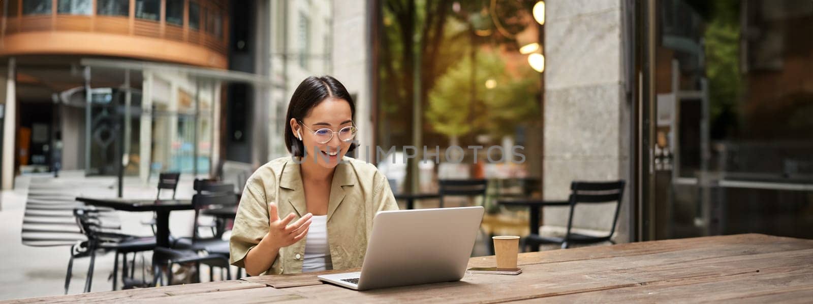 Young woman sitting on online meeting in outdoor cafe, talking to laptop camera, explaining something, drinking coffee by Benzoix