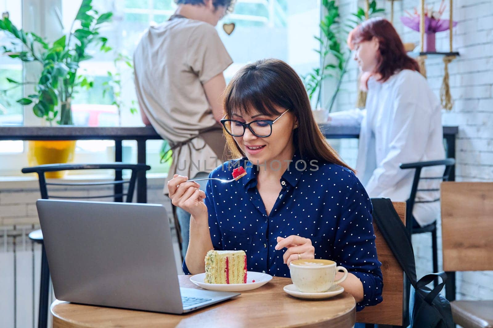 Middle aged woman in bakery cafeteria with cup of coffee and dessert cake, sitting at table, with laptop. Lunch break, food, lifestyle, mature 40s people concept