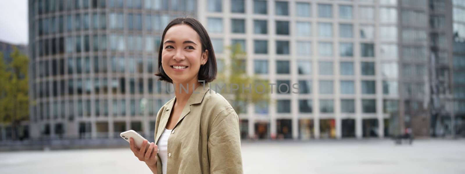 Vertical shot of young asian digital nomad, girl talks on mobile phone and walks on street with laptop. Young woman remote worker going to coworking space by Benzoix