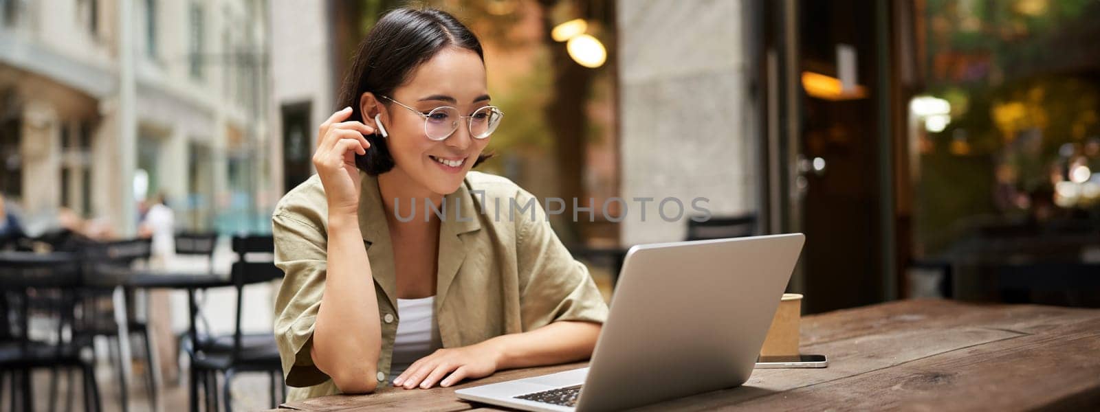 Portrait of asian girl works in cafe, uses laptop and sits outdoors on street. Digital nomad and online learning concept.