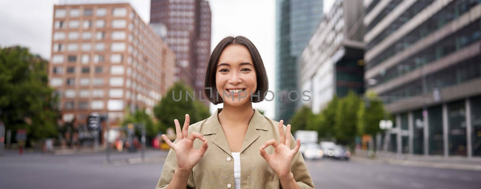 Okay, no problem. Smiling young asian woman shows ok gesture, approve smth, like and agree, accepting, standing on street of city.