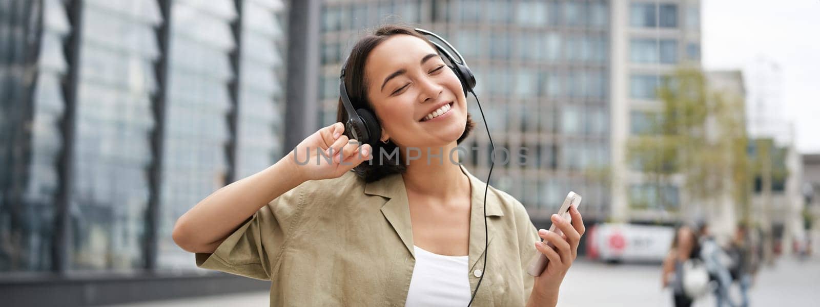 Happy girl dances on street and listens music in wireless headphones, holds mobile phone, using streaming app.