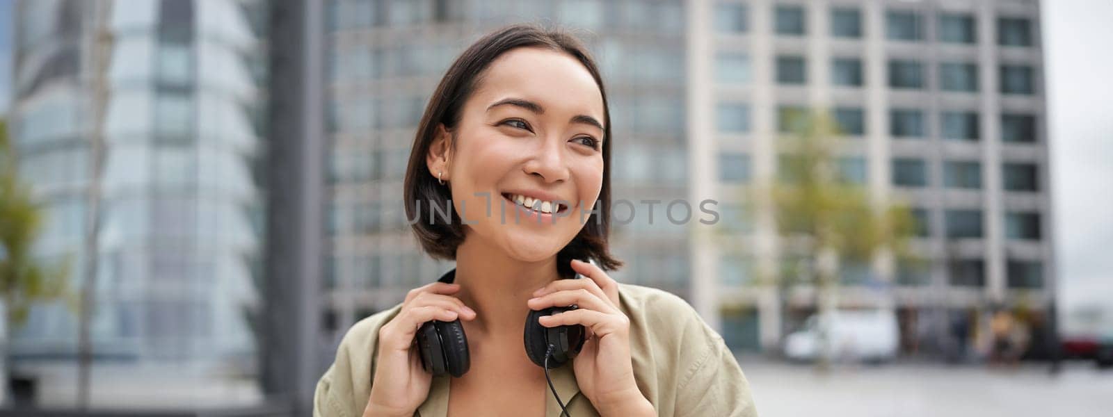 Portrait of smiling asian girl with headphones, posing in city centre, listening music.