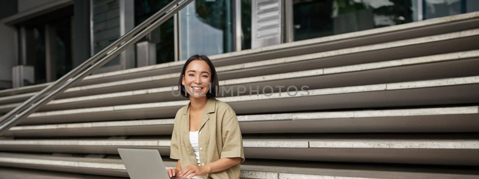 Vertical shot of asian girl sits with laptop, drinks coffee on university stairs. Young woman, student does her homework outdoors by Benzoix