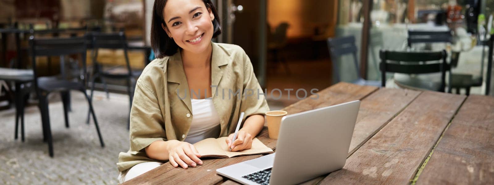 Portrait of young woman sitting in cafe with laptop, making notes, smiling at camera, working, having online meeting.