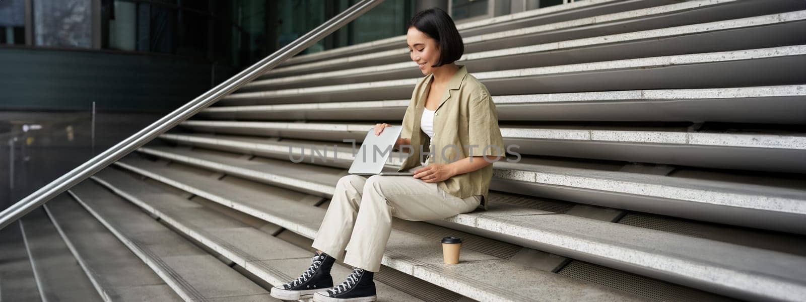 Young smiling asian girl with laptop, sits on street of city with coffee. Young woman does homework on computer while sitting on street stairs, digital nomad works remote by Benzoix