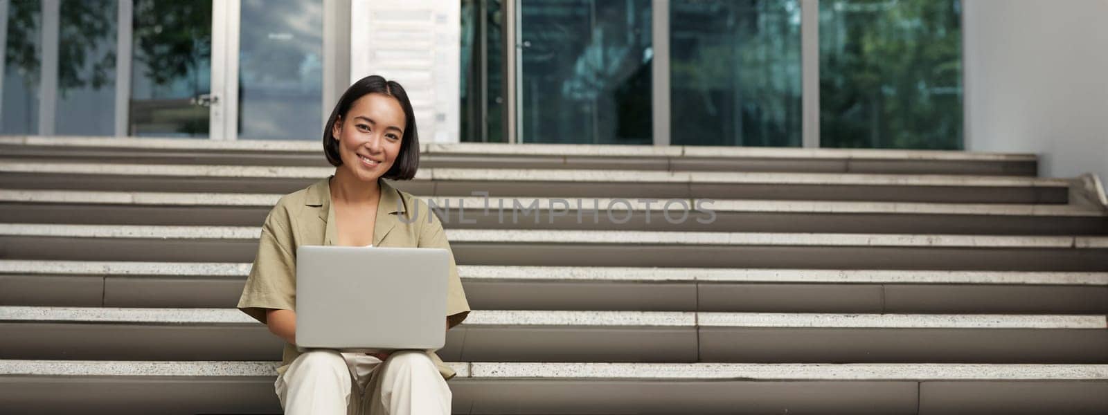 Young asian woman using laptop outdoors. Cute girl student with computer, does her homework, types, sits on stairs outside campus by Benzoix