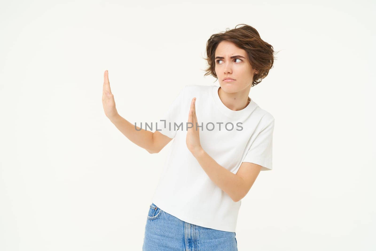 Portrait of woman avoiding someone, shows block hands gesture and refusing from something bad, standing over white background by Benzoix
