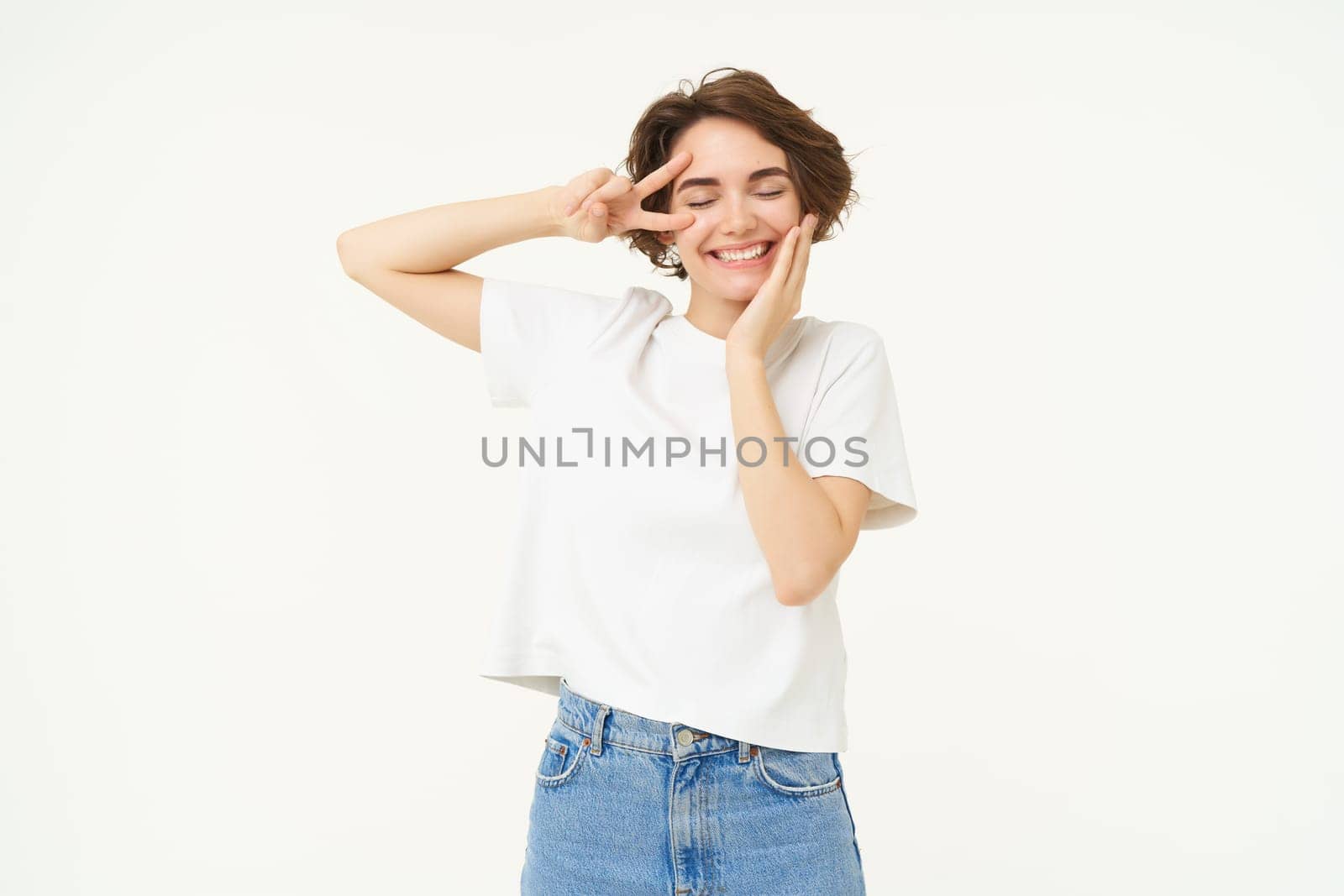 Portrait of young girl shows peace, v-sign and smiling, express posivity and joy, posing over white studio background by Benzoix