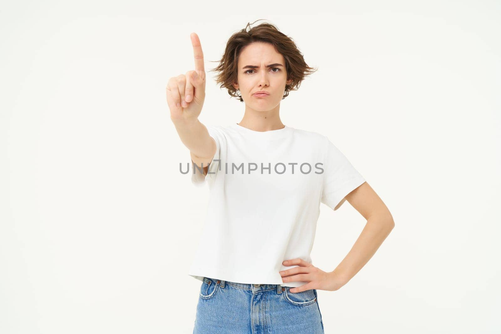 Portrait of serious young woman, shows one finger, stop gesture, disapprove something with frowning face, stands over white background.
