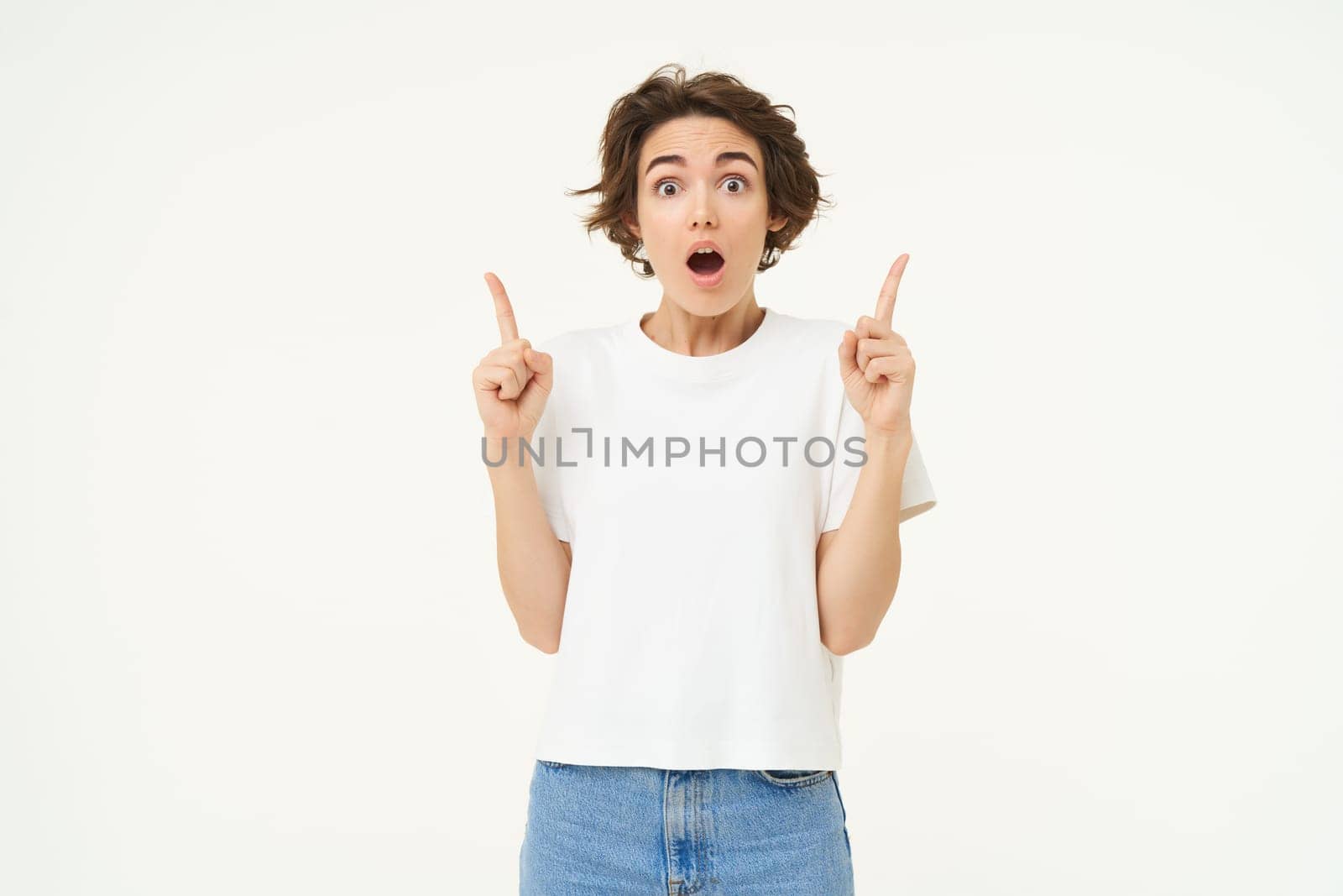 Image of brunette woman pointing fingers up, showing advertisement, gasping and looking amazed, saying wow, impressed by offer, standing over white background.