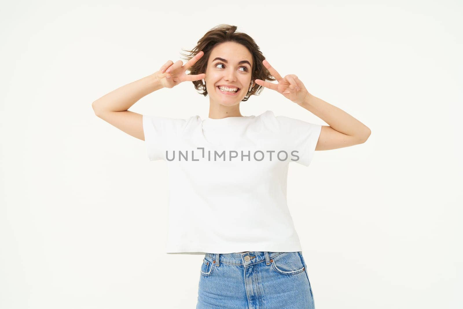 Happy smiling woman, showing peace, v-sign gesture and listening to music in wireless headphones, posing over white background. Copy space