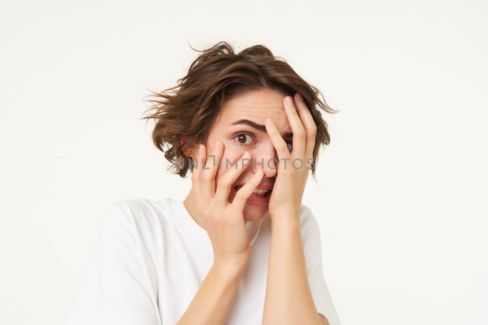 Close up of brunette woman hiding her face, screams and looks frightened, horrified by something, standing scared against white background. Copy space