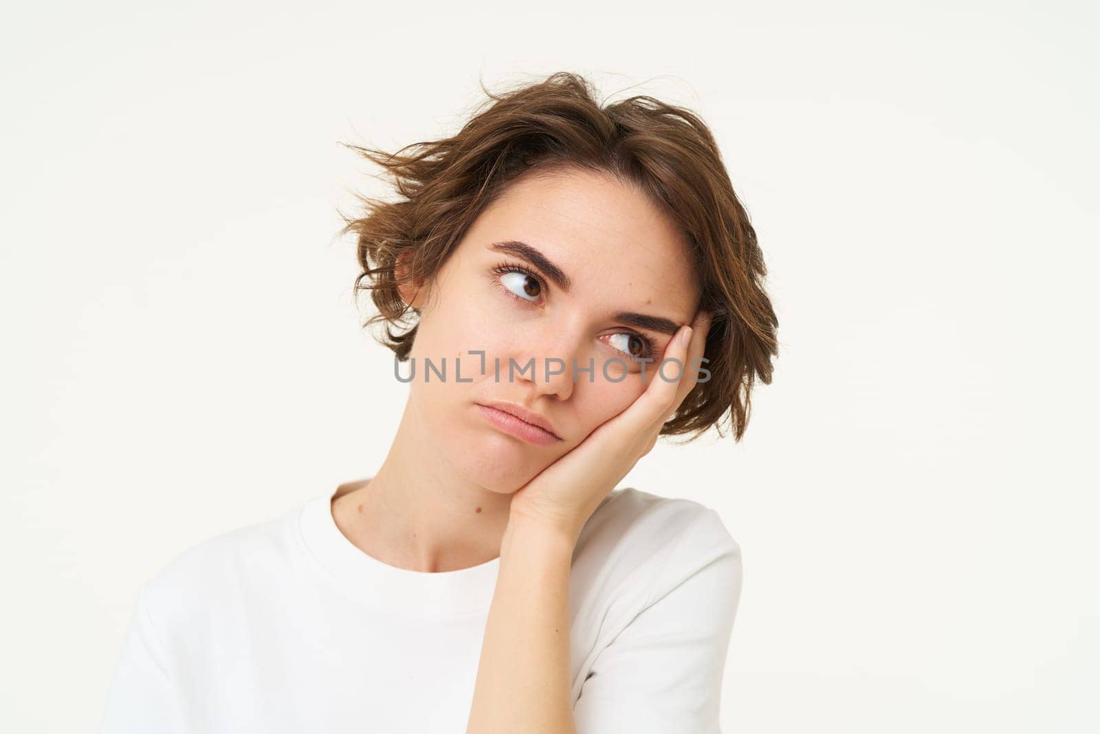 Portrait of young bored woman, leans head on hand and frowns, looks upset and grumpy, thinking, standing over white background.