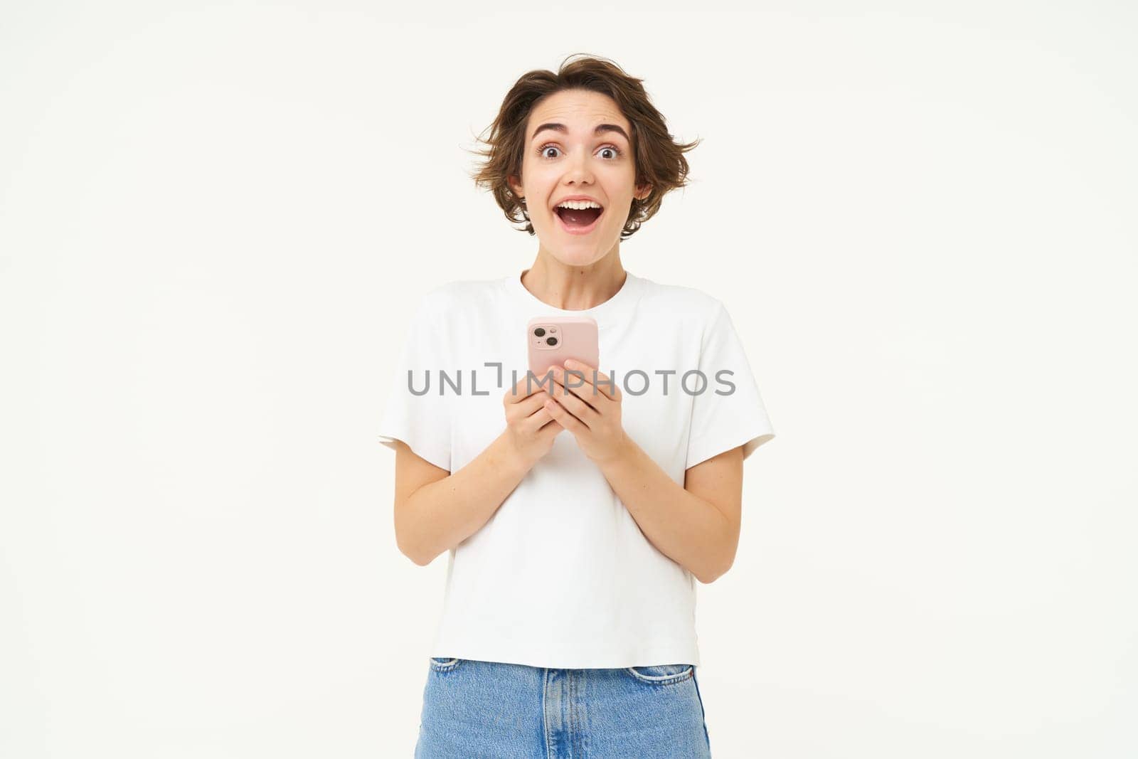 Portrait of brunette girl with shocked face, reading something amazing on mobile phone, standing with smartphone over white background by Benzoix