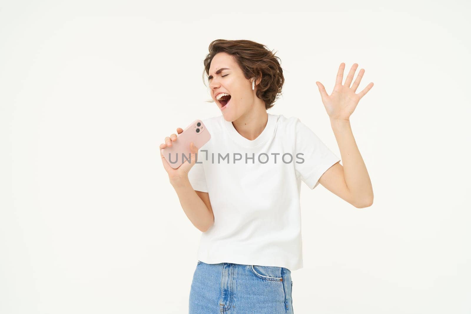 Portrait of happy young woman singing in mobile speakerphone, listening music in wireless headphonse, playing smartphone karaoke game, standing over white background.
