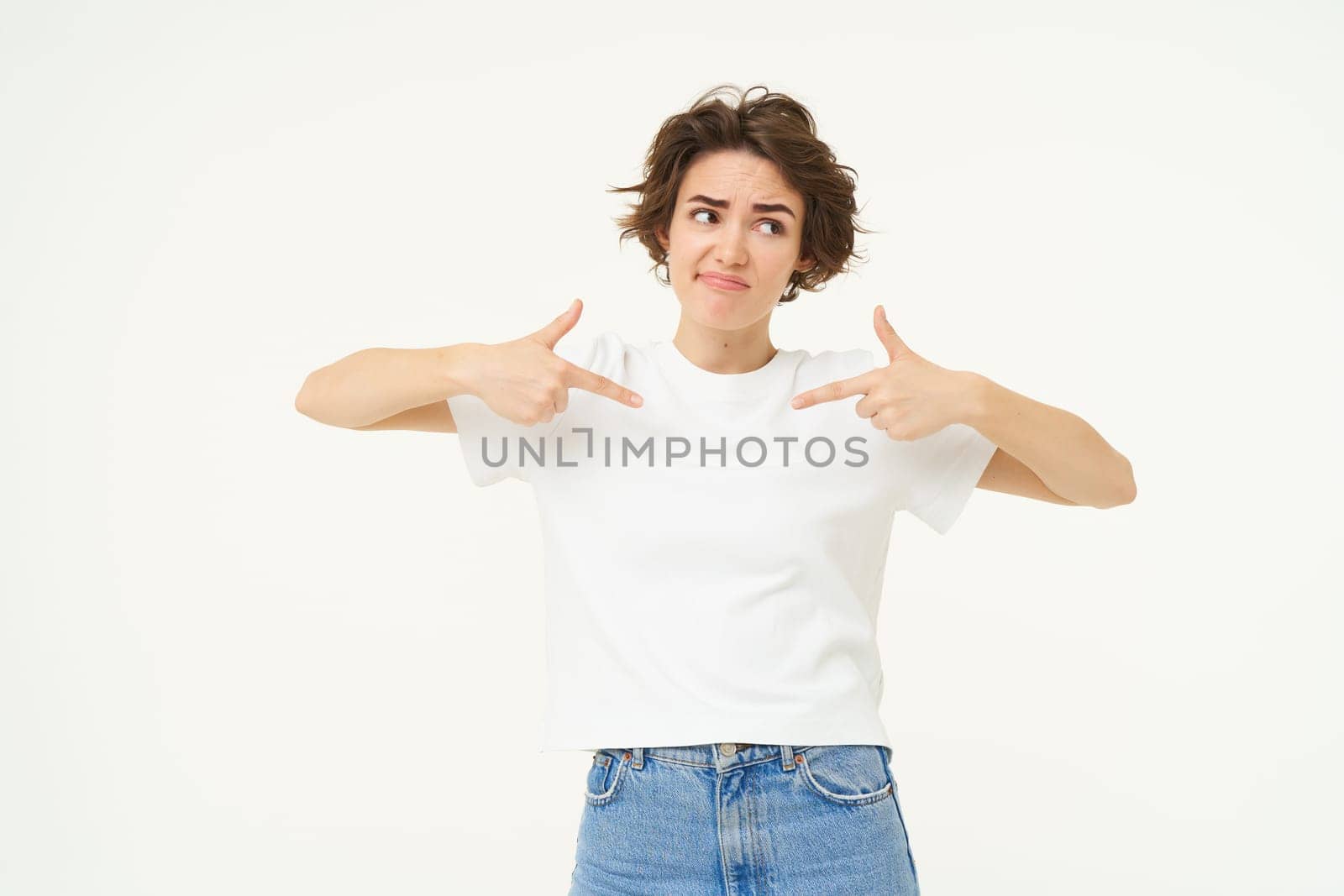 Portrait of unsure brunette girl, pointing at herself and looking uncertain, looking away, standing over white studio background. Copy space