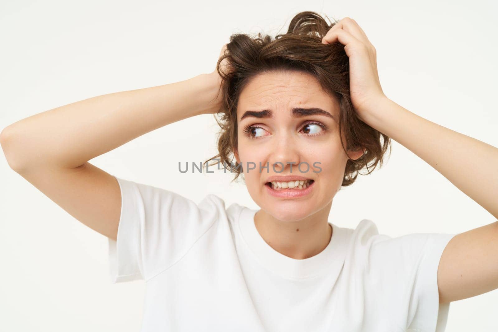 Close up of woman in panic, holding hands on head and clenching teeth, looking troubled, puzzled and frustrated by something, standing over white background.