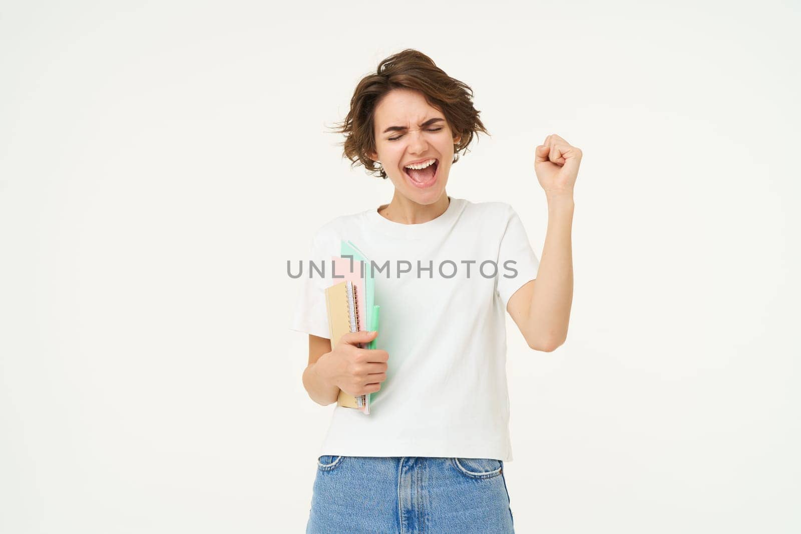 Excited and enthusiastic girl, feeling thrilled, makes fist pump and screams from joy, posing with notebook against white studio background.