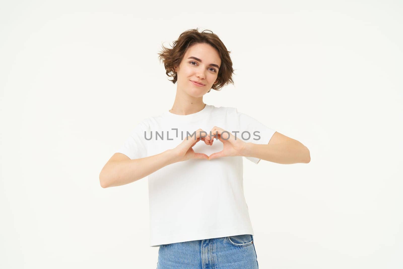 Portrait of brunette girl shows heart sign, love gesture, express care and warm feelings, stands over white background.