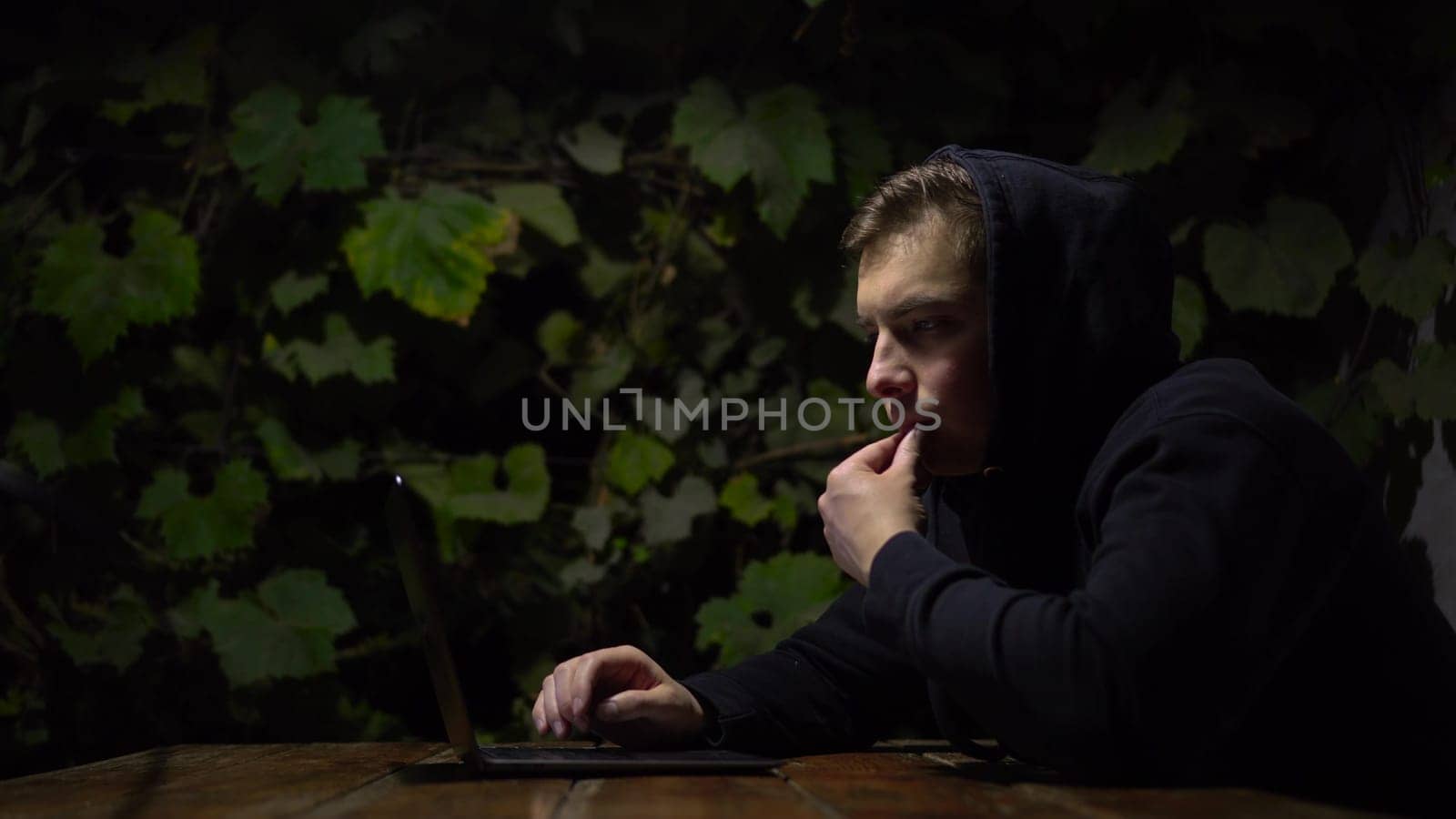 A young man at a table in a gazebo works on a laptop during late at night. A man in a black hoodie with a laptop against a background of green grapes. 4k