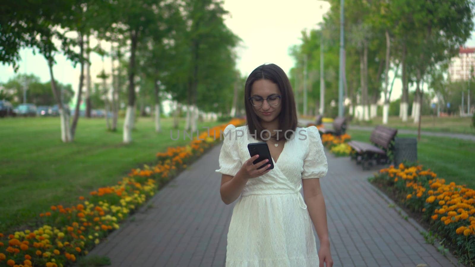 A young woman walks along a flower alley with a phone in her hands. A girl in a white dress looks at her phone and walks among the flowers. 4k