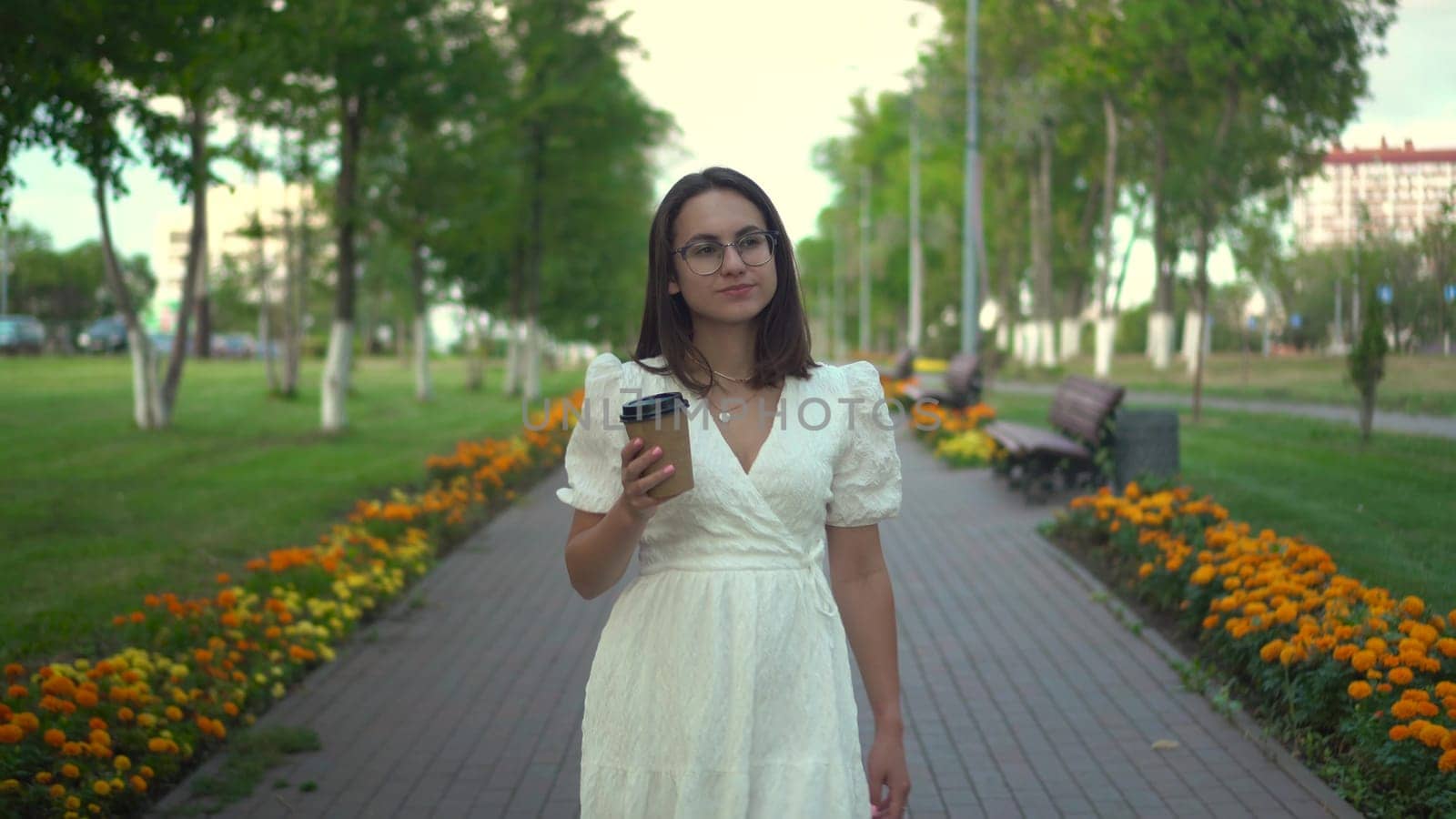 A young woman walks along an alley in a white dress and drinks crofe. A girl with glasses walks through the park with coffee in her hands. 4k
