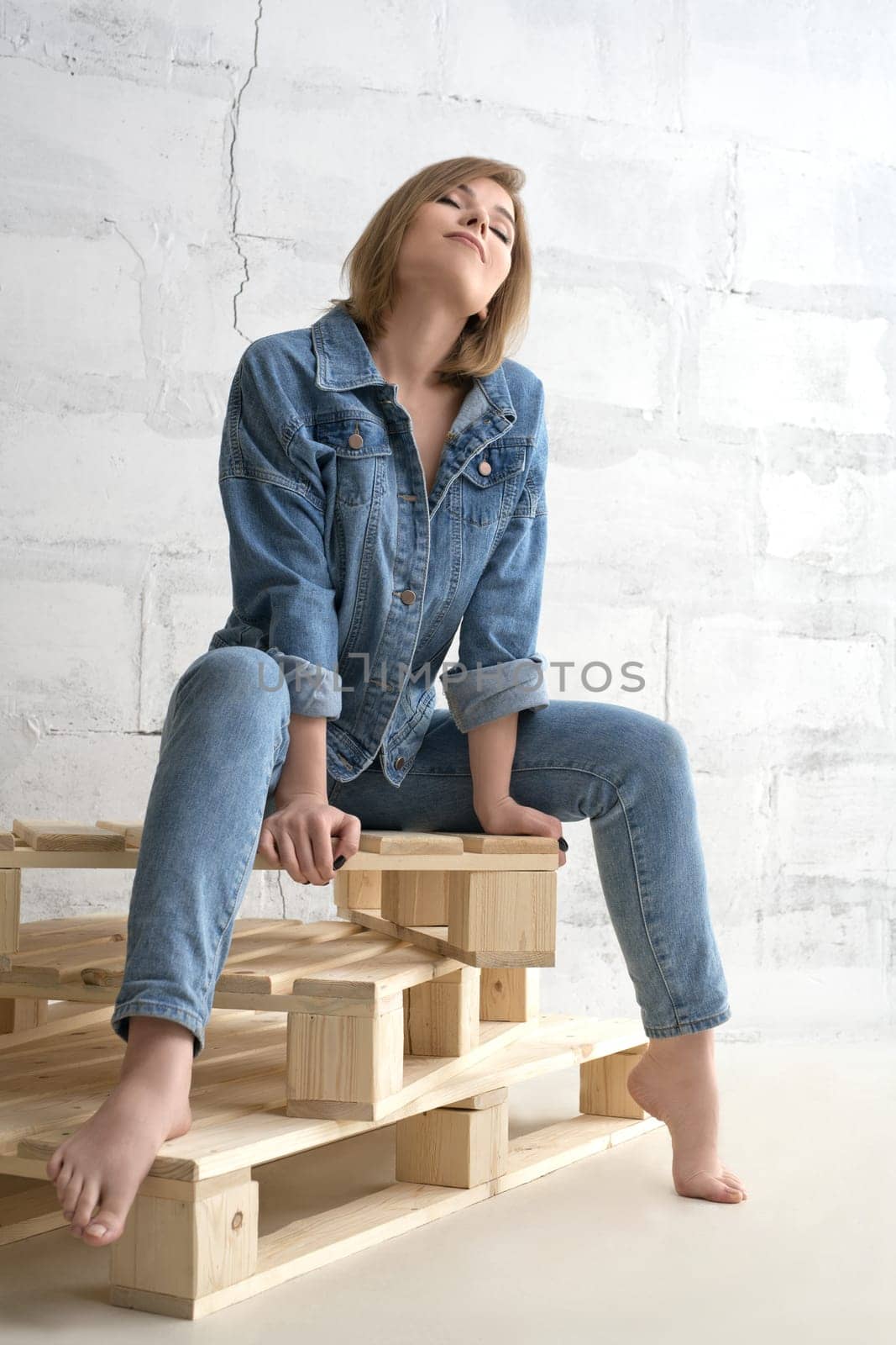 Pretty sexy girl in jeanswear sitting on wooden pallet shot against brick wall