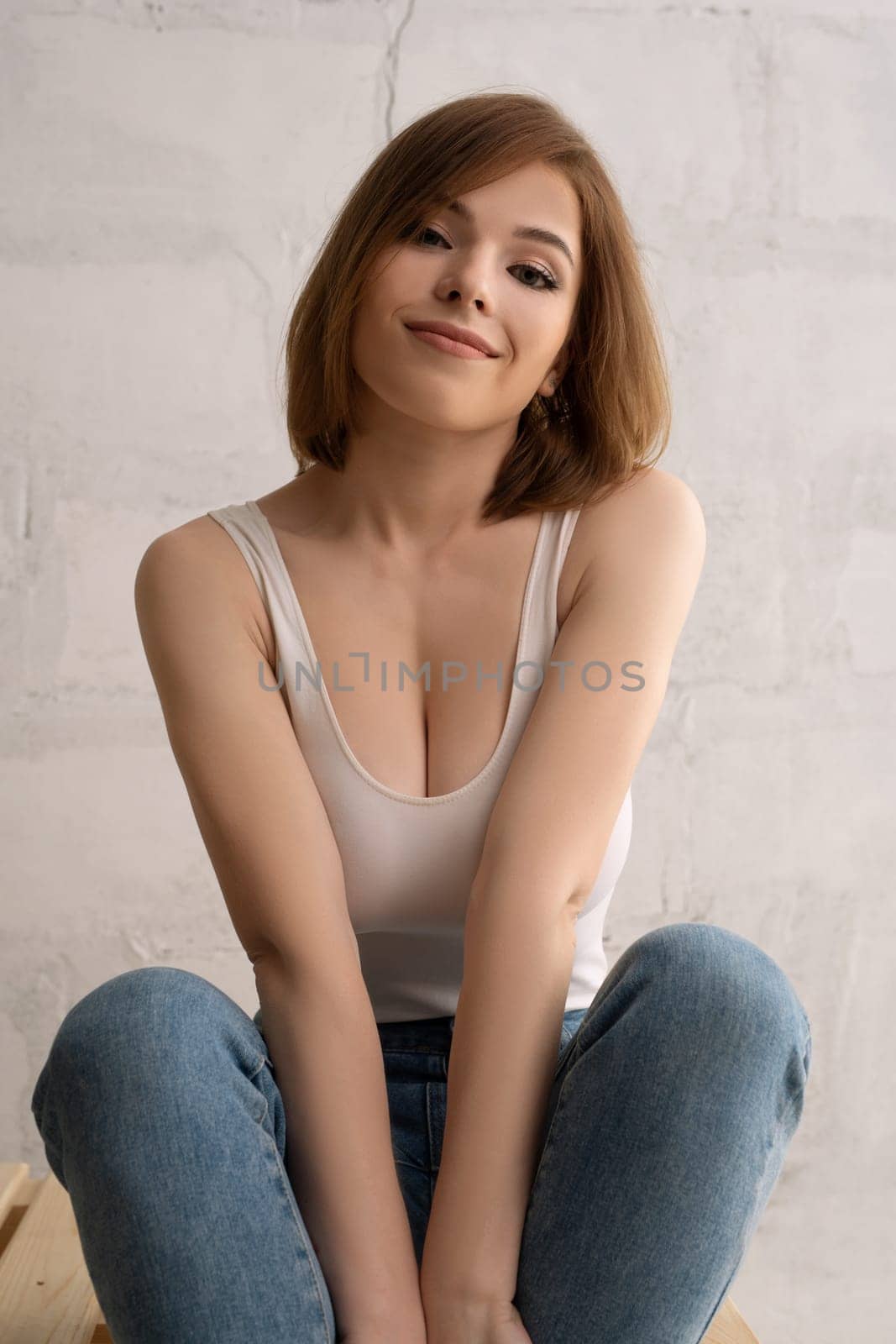 Young happy brunette wearing white top and jeans sitting on her knees against brick wall
