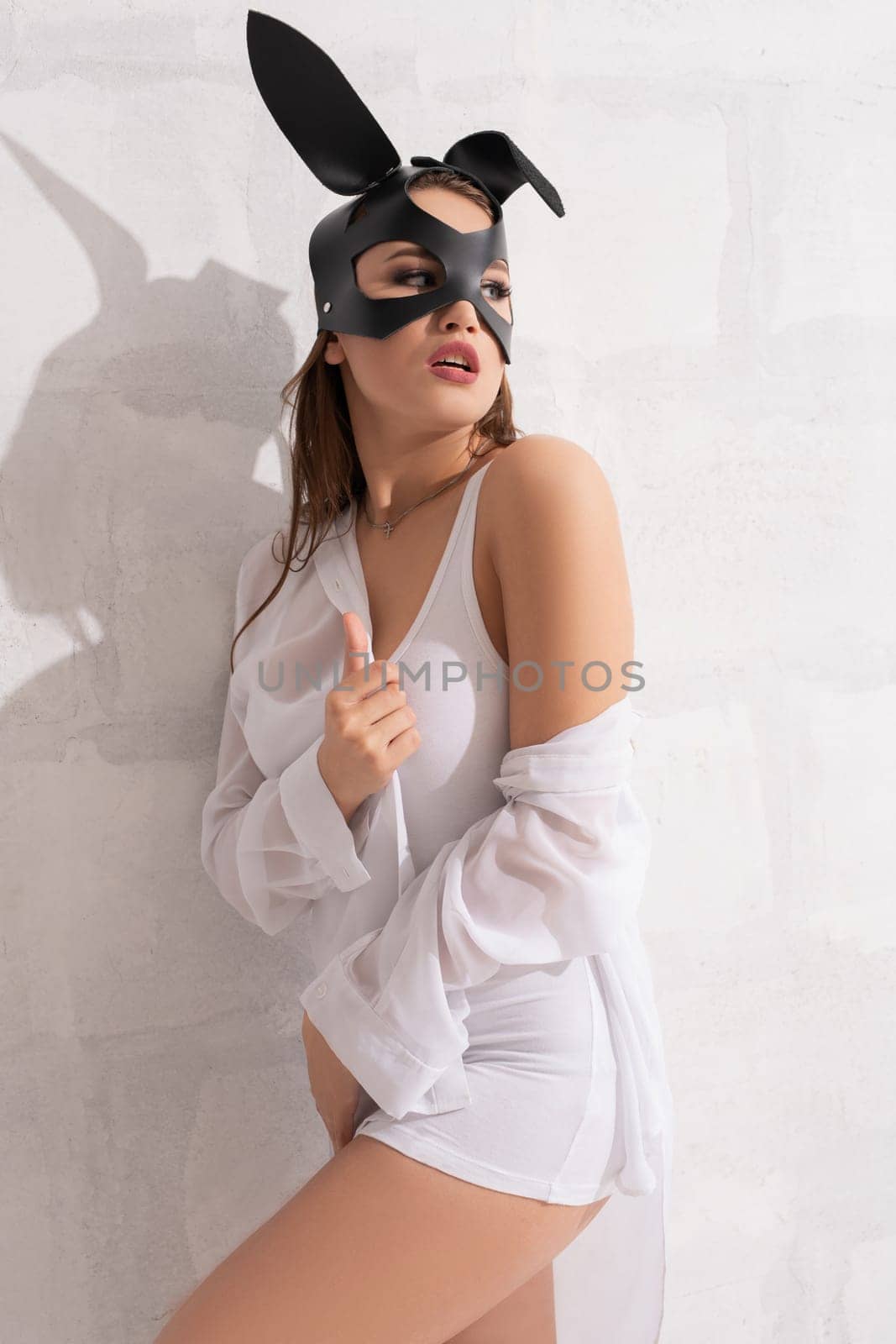 Woman in a bdsm black leather bunny ears mask wearing only a shirt view against brick wall