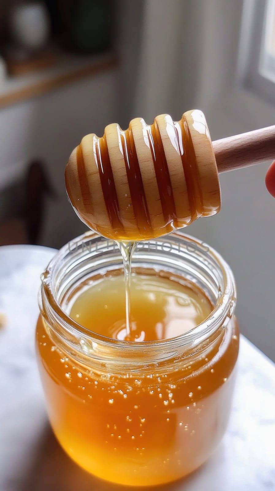 Golden honey is dripping from a wooden honey dipper into a glass jar, creating small bubbles on the surface
