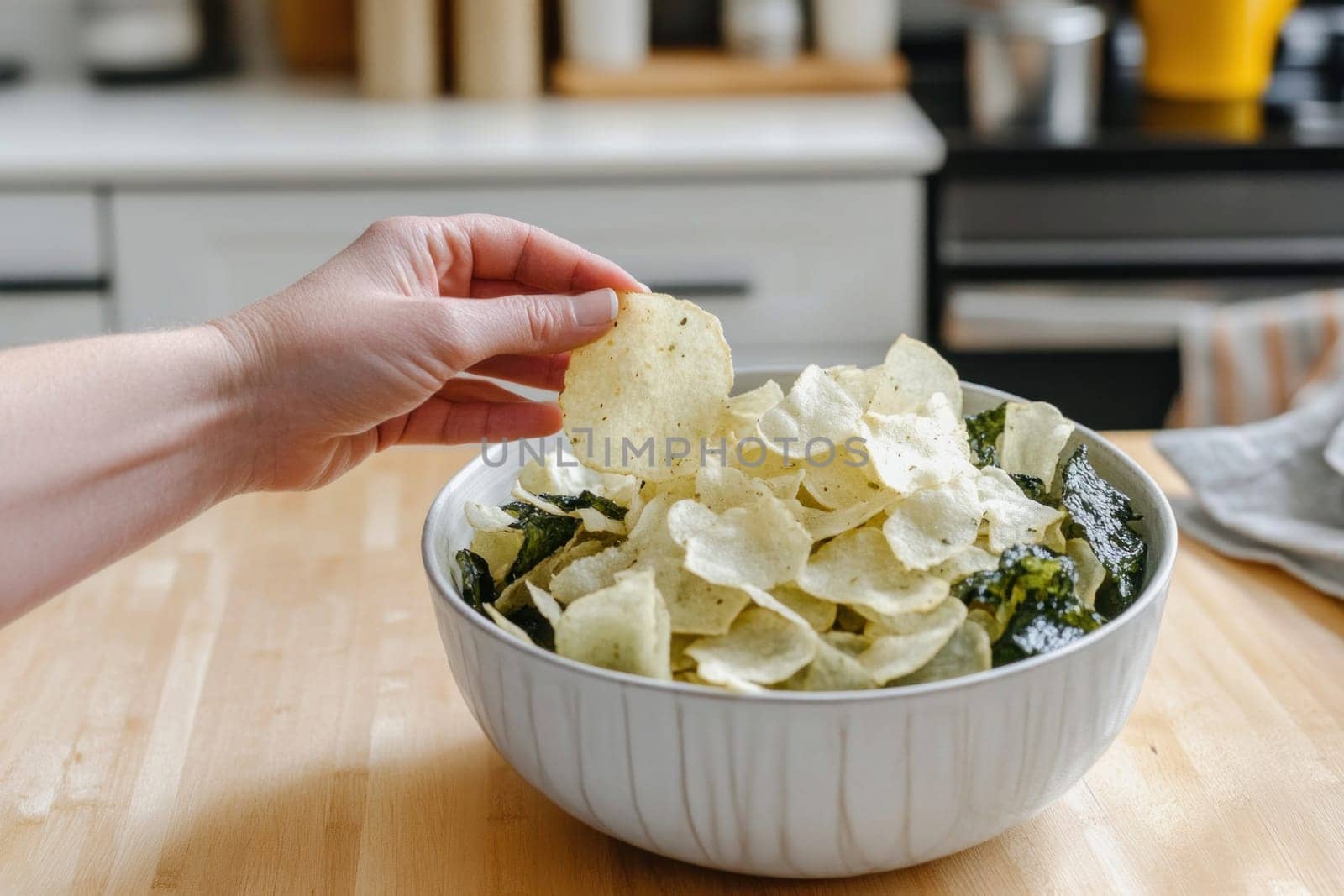 Hand grabbing a seasoned potato chip from a bowl filled with chips and seaweed snacks in a bright kitchen