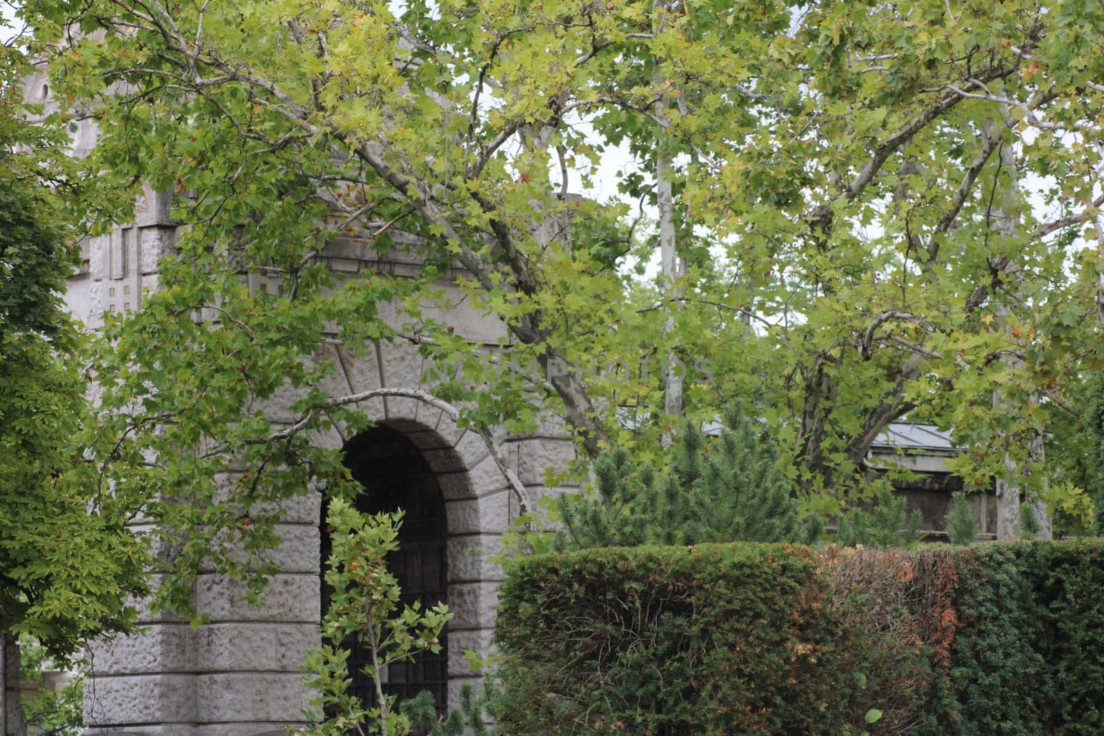 The historic stone archway stands amidst vibrant green trees and bushes, creating a peaceful atmosphere in the park as clouds drift overhead.