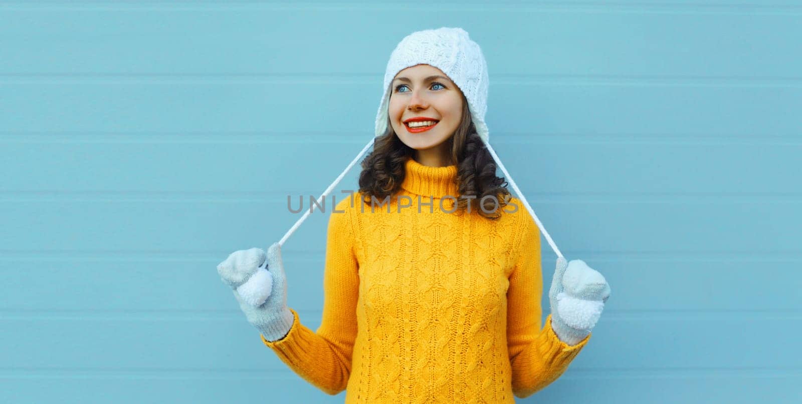 Winter christmas portrait of happy young woman smiling in yellow sweater, white hat looking away standing on blue background