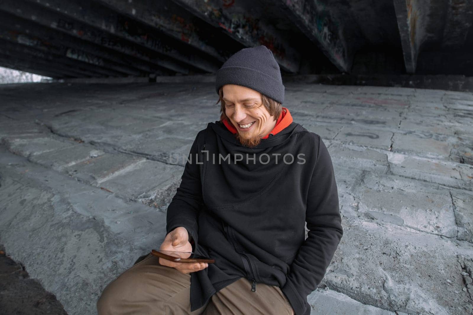 A cheerful individual sits on a concrete surface beneath a bridge, engrossed in their smartphone, smiling and enjoying the simple pleasure of connecting with others.