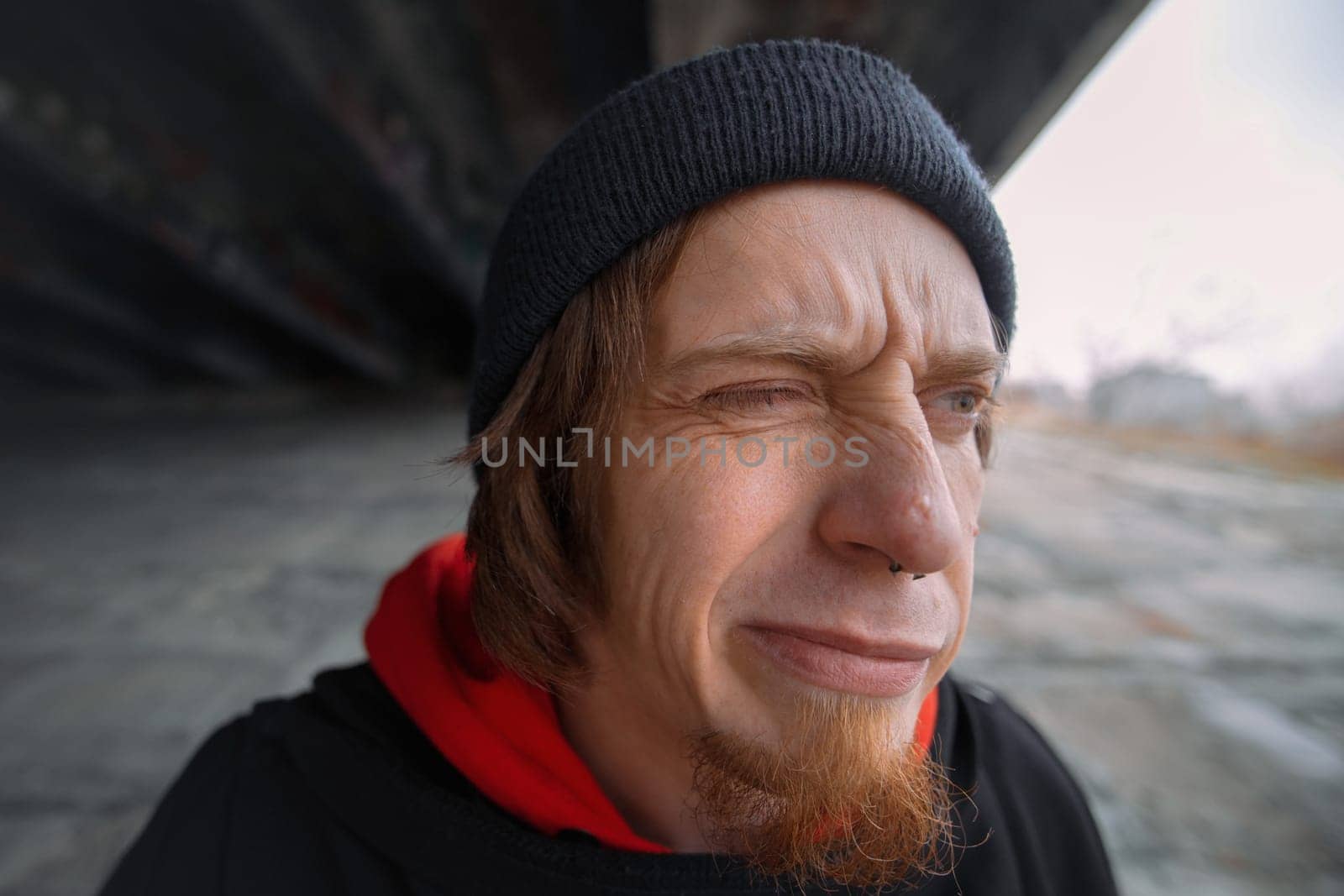Beneath a sturdy bridge, a young man sports a beanie and hoodie, squinting slightly while lost in thought, the warm light illuminating his features.