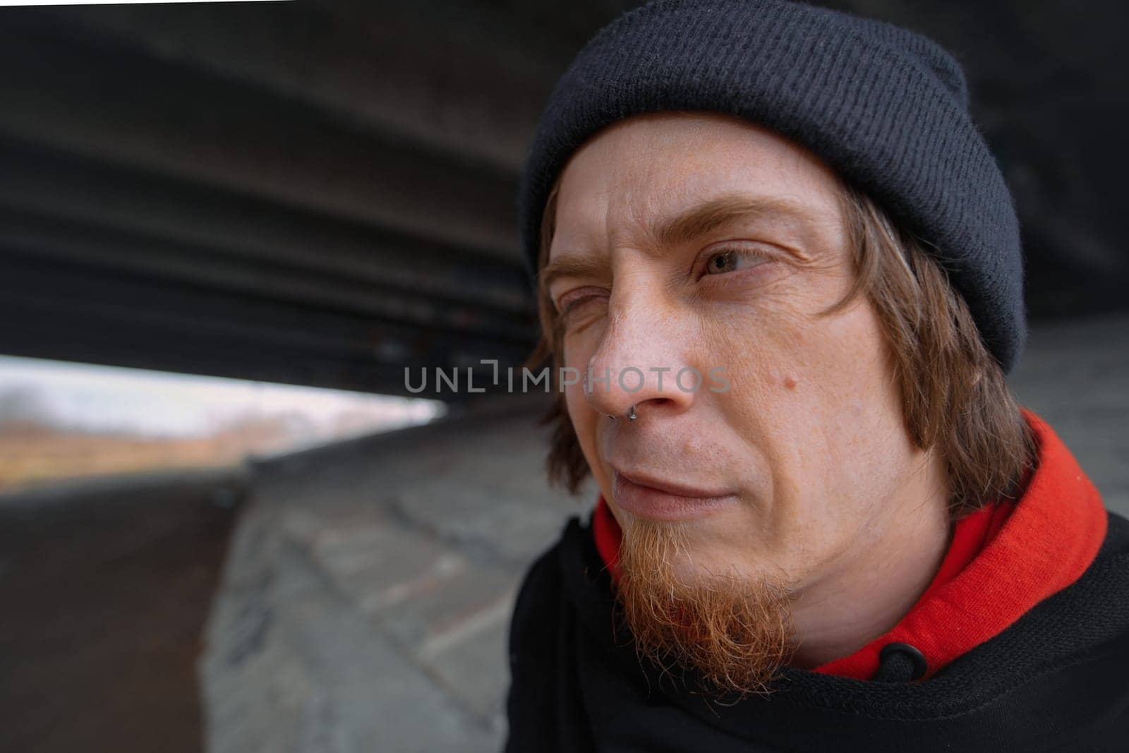 Caught in a quirky moment beneath the bridge, a man sporting a beanie and a red hoodie flashes a playful expression, adding character to the urban setting.