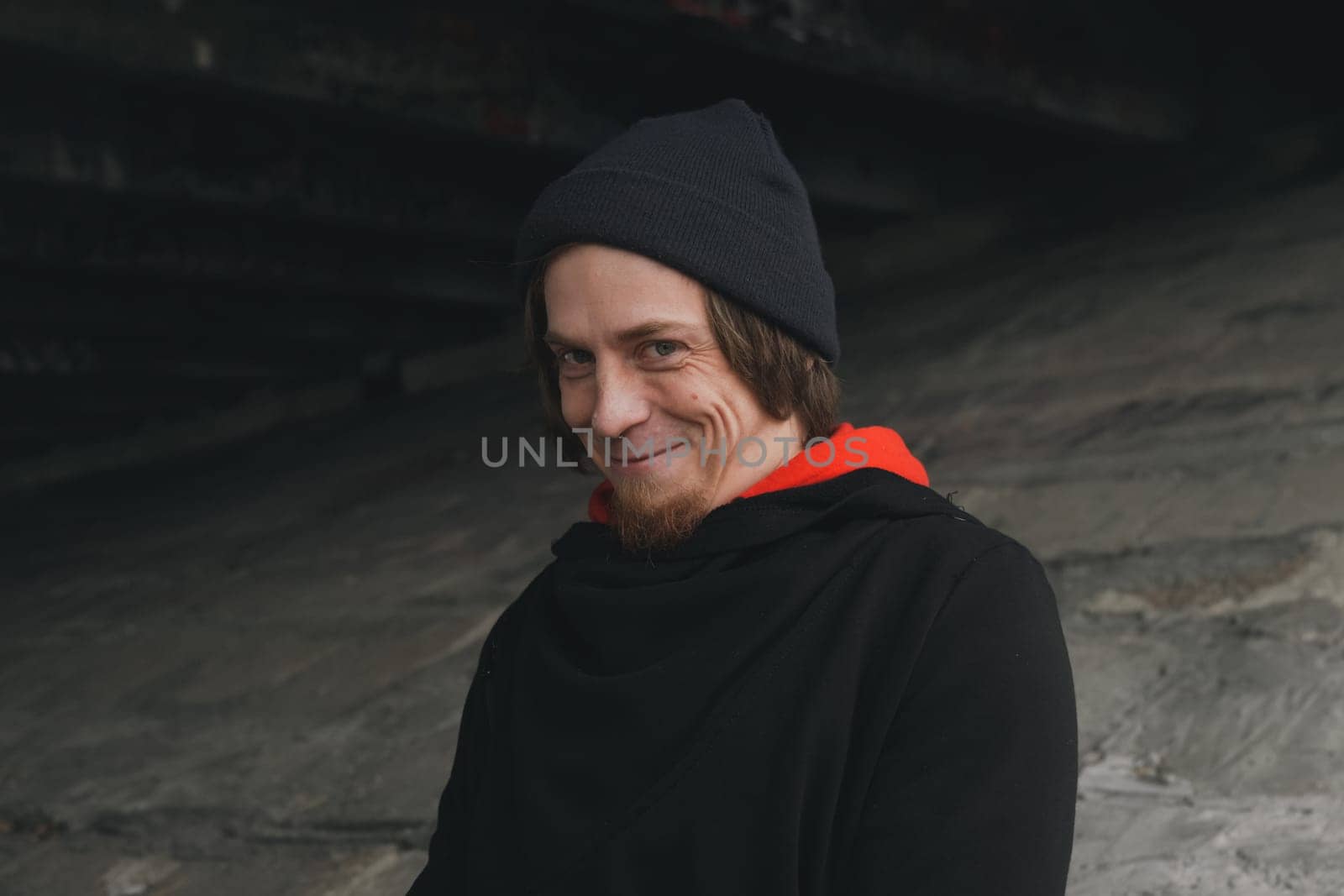 A man with long hair and a beard sits quietly under a bridge, lost in thought, wearing a beanie and hoodie. The urban setting evokes calm despite the bustling city above.