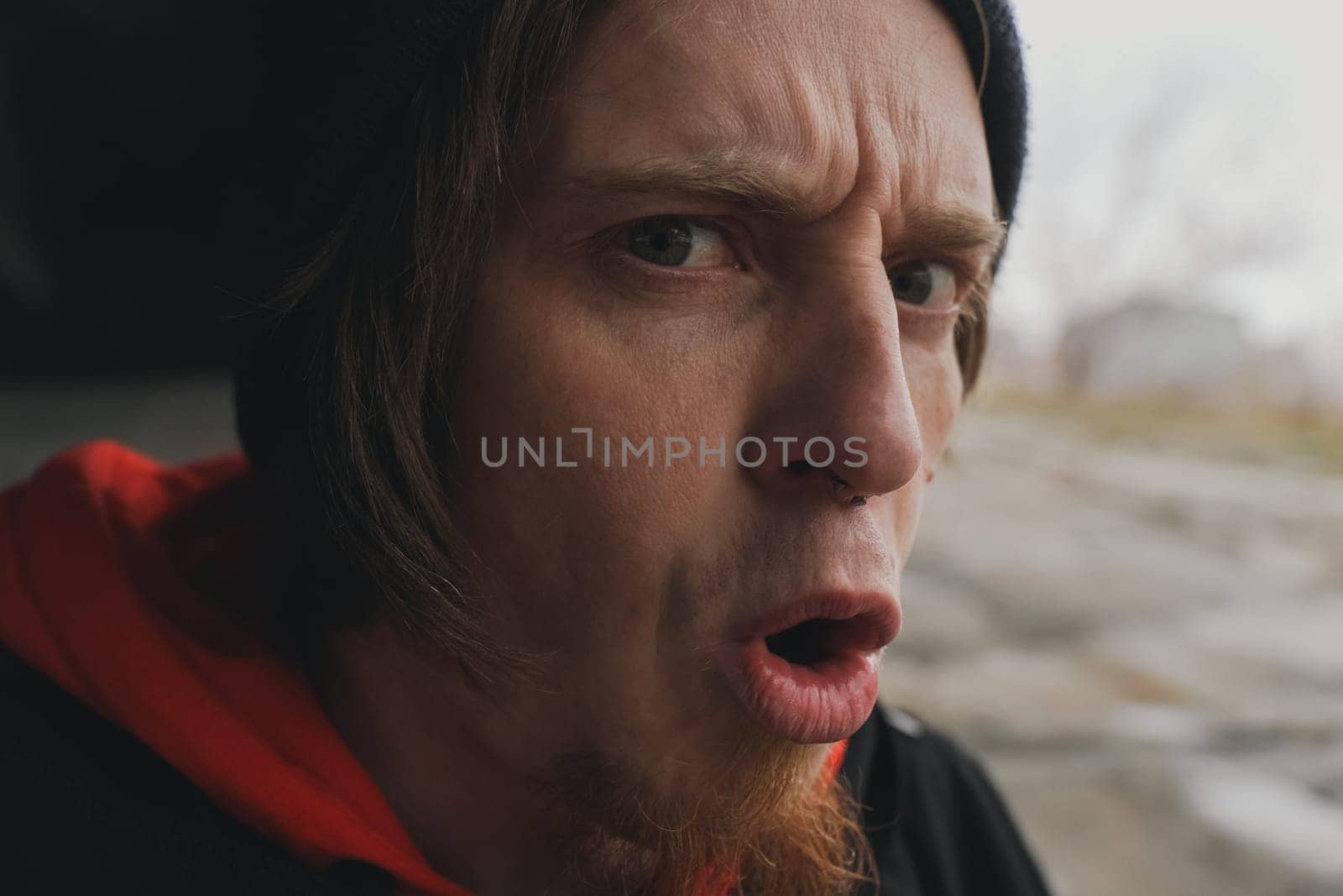 Expressing astonishment, a man with a beard wearing a hooded sweatshirt reacts to something unexpected in a deserted location on an overcast day.