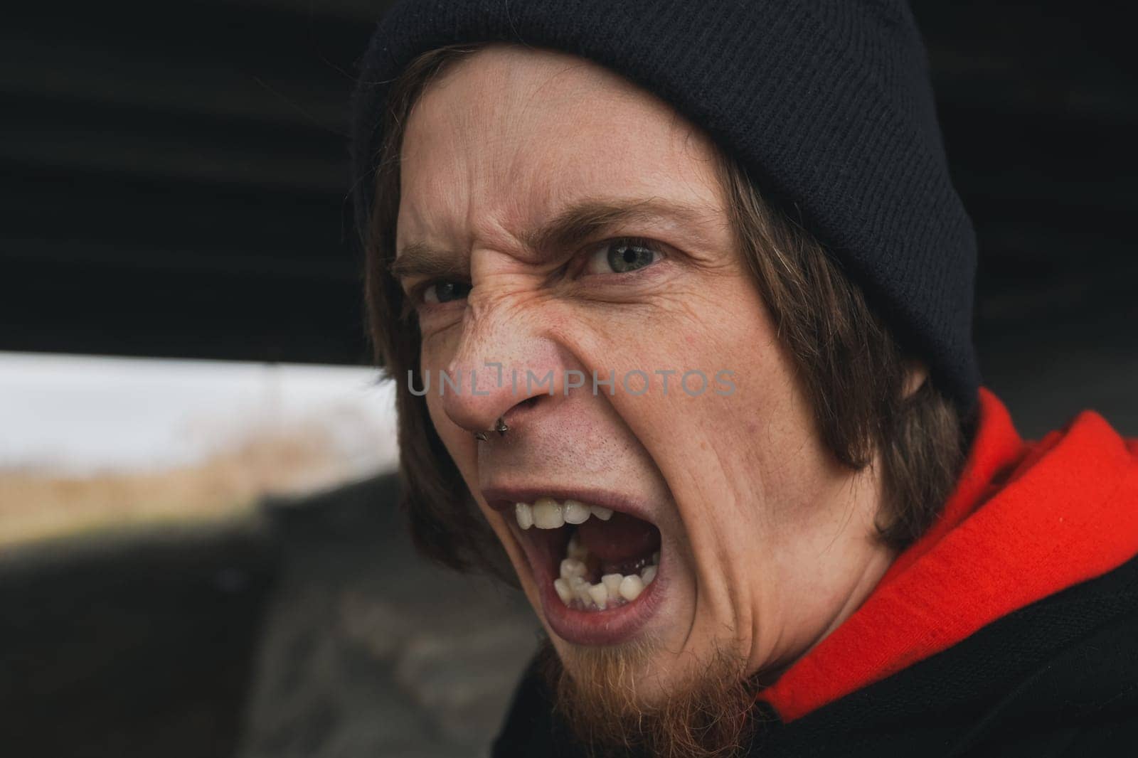 A young man, wearing a black beanie and a vibrant red hoodie, is caught in a moment of raw emotion, shouting fervently beneath a bridge.