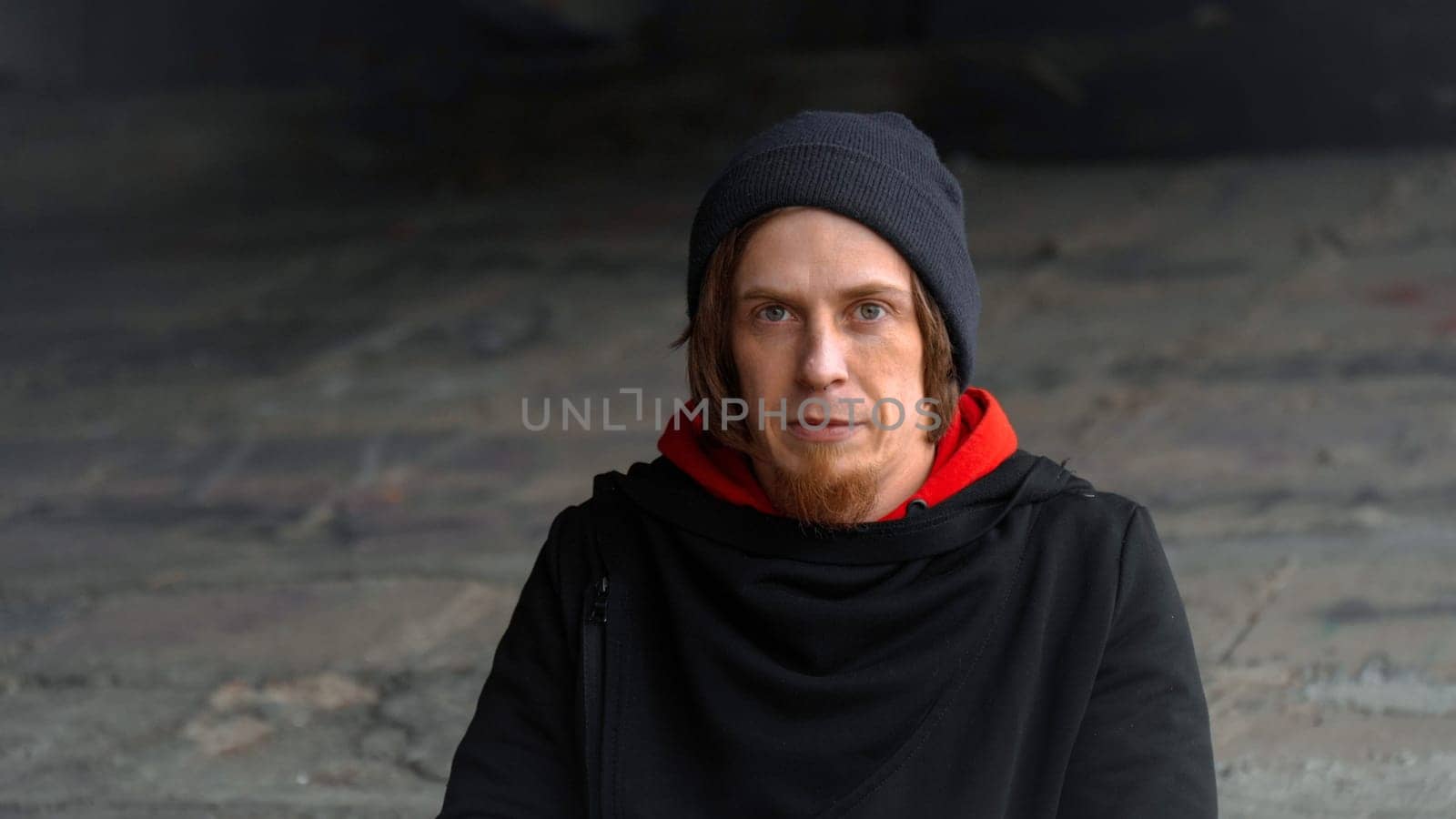 In a dimly lit urban space, a man gazes thoughtfully while wearing a red hoodie and a beanie.