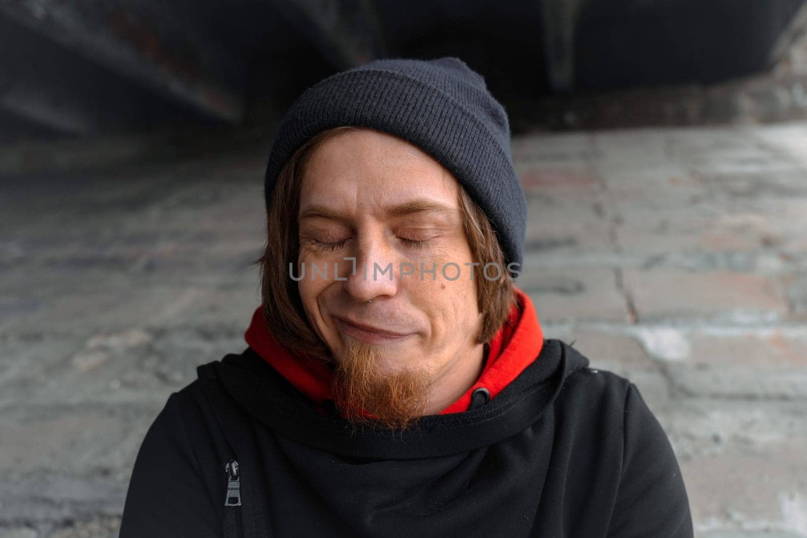 A man with long hair and a beard sits quietly under a bridge, lost in thought, wearing a beanie and hoodie. The urban setting evokes calm despite the bustling city above.