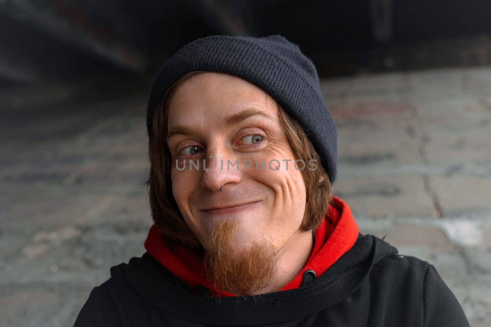 A man with long hair and a beard sits quietly under a bridge, lost in thought, wearing a beanie and hoodie. The urban setting evokes calm despite the bustling city above.